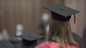 Video from a backside of a young girl who is graduated from the uni. She is dressed in a black and red mantle and square academic cap. She is standing in a big hall with all of her coursemates.