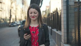 Gorgeous Ukrainian girl walking through  street with smartphone in hand. Nice smiling young woman wearing black leather jacket. Spring time. Outdoors, sms, social, student, adult