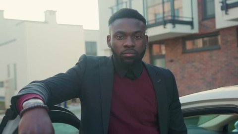 Handsome african american man stand near white car look at camera sunset young vehicle open passenger arm auto street automobile businessman buy jacket portrait close up slow motion