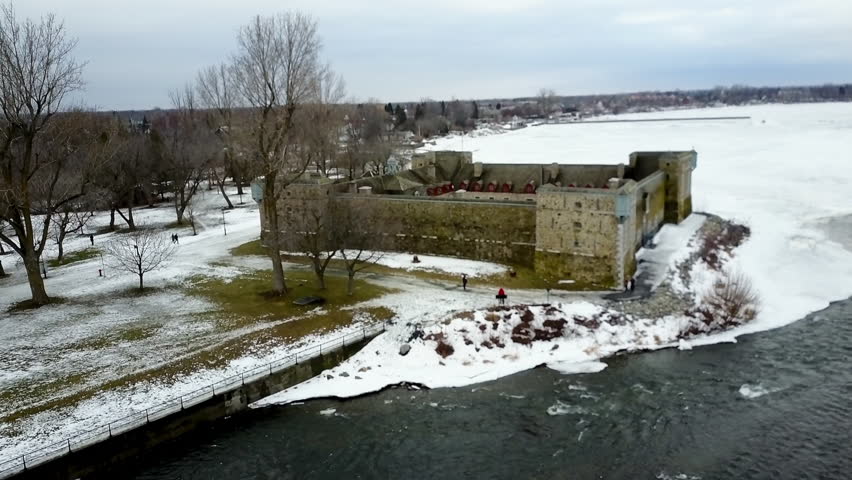 Cinematic aerial / drone footage ascending over the lake with dry trees, snow, houses and a fort in the background in Chambly, Quebec, Canada during winter season.