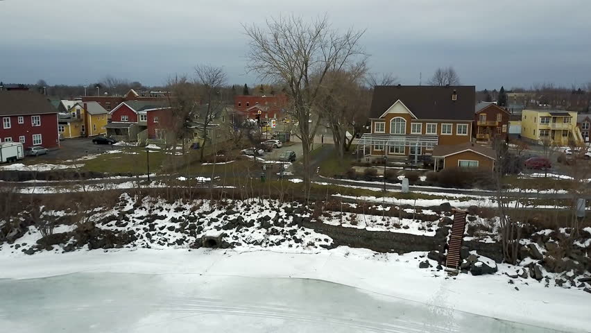 Cinematic aerial - drone footage moving sideways over the lake with dry trees, snow, houses, cars, a walkway in the background in Chambly, Quebec, Canada during winter season.