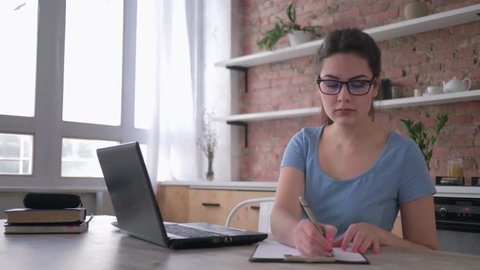 student girl in eyeglasses learning online with computer laptop and writes notes in clipboard sitting at the table in the kitchen