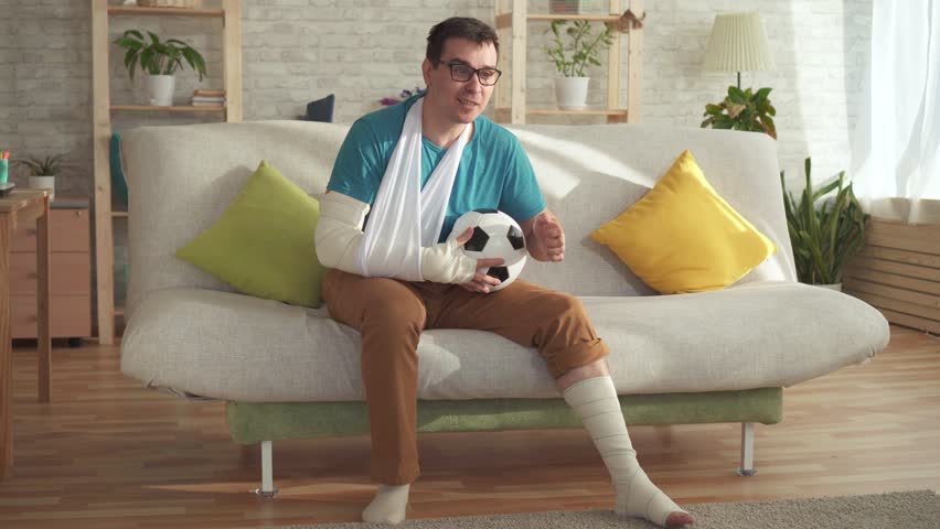 Expressive young man with a broken arm and leg sitting on the couch with a soccer ball learns about win