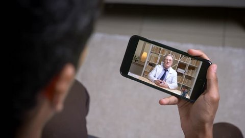 Online videocall via smartphone of senior doctor in white lab coat talking seriously to his patient behind the screen.