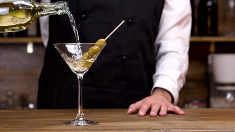 Bartender pouring martini in a cocktail glass in slow motion. Alcohol slowly pours on green olives in the glass.
