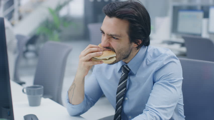 In the Bright Modern Office Young Businessman Eats Hamburger Sitting at His Desktop Computer, He Bites Angrily into the Bun and Continues to Work During His Lunch. In the Background Colleagues Working | Shutterstock HD Video #1027693292