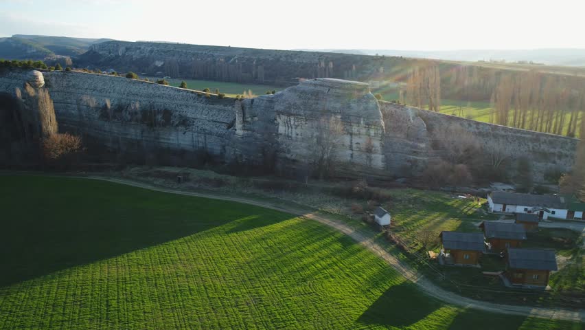 Aerial view of green field and small houses near mountain slope against blue sky. Footage. Beautiful summer landscape
