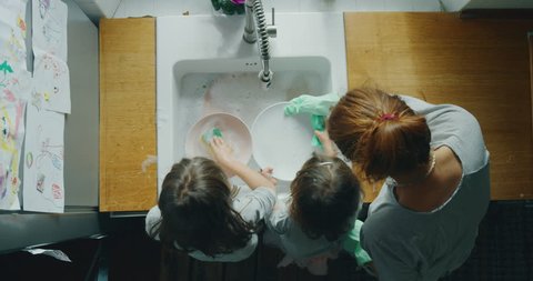 Cinematic authentic shot of little girls sisters daughters having fun to help their mother washing plates in a kitchen at home on a sunny day.