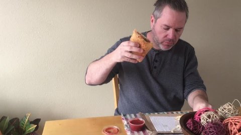 Man eating cheesy calzone lunch at table in daylight after dipping food in marinara sauce. 