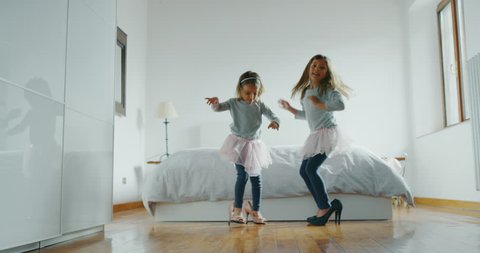 Authentic shot of two little girls in pink tutu skirts are having fun wearing mother high heeled shoes in parents bedroom.