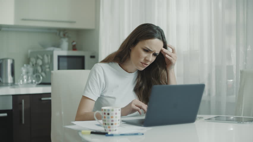 Sad woman working laptop computer at kitchen. Unhappy person use computer technology at home. Worried girl stress at freelance work at home workplace. Upset woman looking at laptop screen | Shutterstock HD Video #1027710653