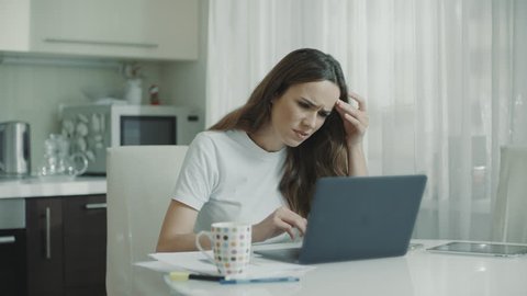 Sad woman working laptop computer at kitchen. Unhappy person use computer technology at home. Worried girl stress at freelance work at home workplace. Upset woman looking at laptop screen