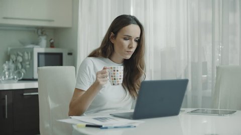 Young woman using laptop computer at kitchen. Portrait of business woman watching news at laptop screen and drinking coffee at home workplace. Female professional working notebook at home
