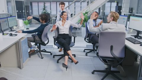 Cheerful Employee Pushes His Beautiful Female Colleague on a Chair Between Rows of Desks with Diverse Business People Working on Desktop Computers in Modern Office Space. Three High Fives Given.