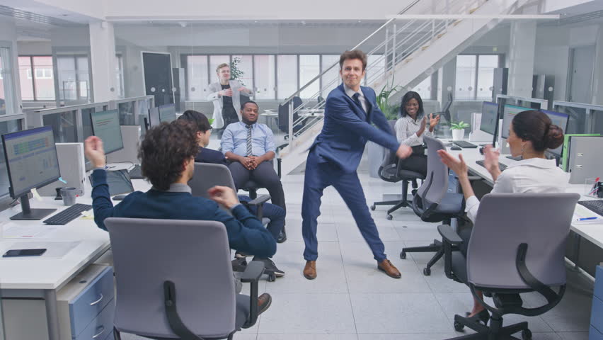 Young Happy Business Manager Wearing a Suit and Tie Dancing in the Office. Colleagues are Cheering. Diverse and Motivated Business People Work on Computers in Modern Open Office. | Shutterstock HD Video #1027712576