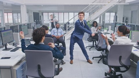 Young Happy Business Manager Wearing a Suit and Tie Dancing in the Office. Colleagues are Cheering. Diverse and Motivated Business People Work on Computers in Modern Open Office.