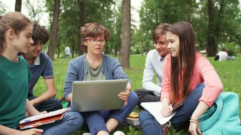 Panning of Caucasian teenagers gathered around their female teacher sitting on lawn with laptop on her knees and explaining new lesson theme