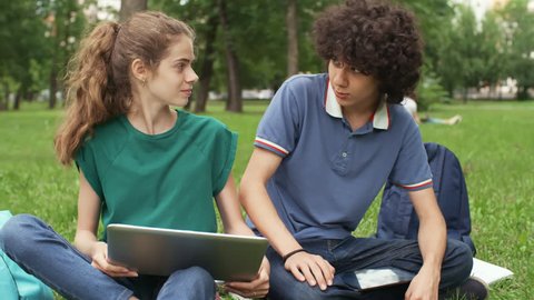 Panning of Caucasian schoolgirl and schoolboy sitting on lawn with laptop and discussing home task together
