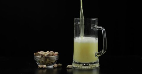 Beer is pouring into glass on black background. Bowl of pistachios nuts.