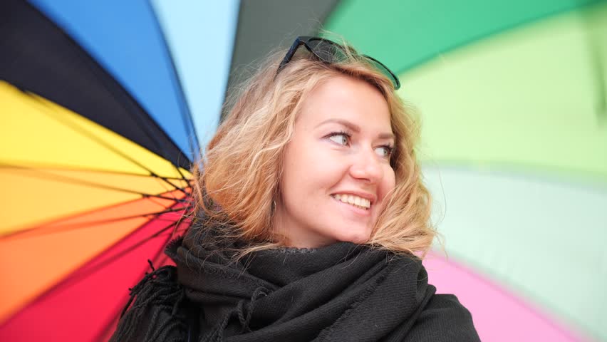 Young beautiful blonde girl in black scarf smiles happily and twists her big colorful umbrella.
