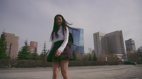Pretty Thai student turns around whole body, standing in front of great city. From bottom angle you can see her slender legs. Woman turns around gracefully, and wind slightly touches her short skirt.