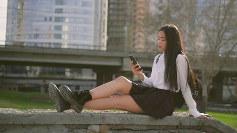 Chinese cute girl is chating on cell phone. Female is nodding in likely pose. Shi's in white shirt and short skirt sitting on rock and being serious. City near is large. Wind blowing her hair nicely.