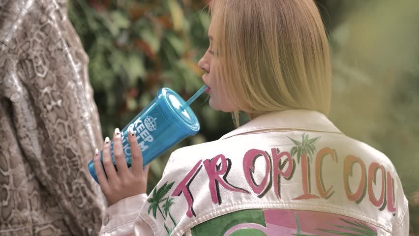 A blonde girl sits with her back to the camera and drinks lemonade or a cocktail from a blue tube. She has a blue glass in her hand, and another girl is standing next to her in a leopard jacket.