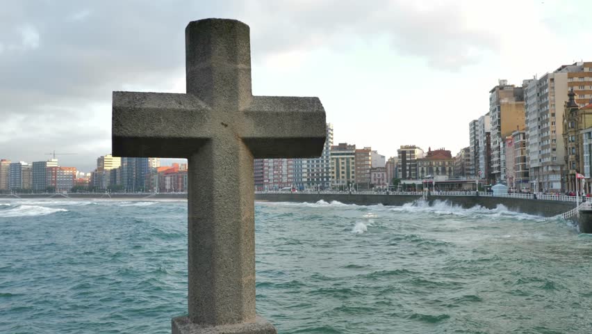 A big stone cross in front of the skyline from the city of Gijon (Asturias, Spain); tall buildings facing the sea, waves, a cool mood.