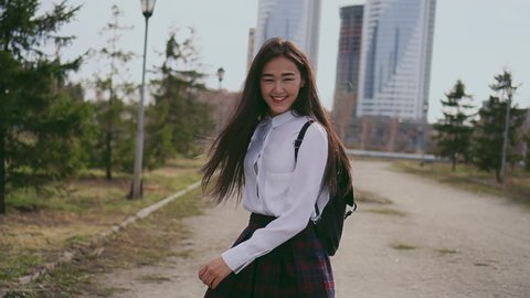 Smiling japanese schoolgirl in short skirt jumping along the street after classes. Girl looks straight into camera, laughs and rejoices life, turns around and jumps again for happiness to skyscrapers.