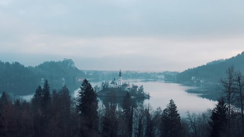 AERIAL: A drone shot flying over the forest and approaching an island on Lake Bled in the middle of the cold winter morning. Famous tourist destination.