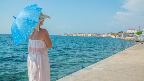 PIRAN, SLOVENIA - 7. AUGUST 2018 A young woman is standing on the pier and she is is holding a blue umbrella in her hands and is also wearing a nice straw hat.