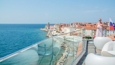 A nice restaurant is overlooking a sea town of Piran. A young woman is standing on the other side of the balcony. It's a nice summer day.
