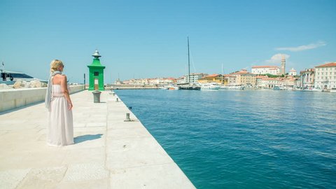 PIRAN, SLOVENIA - 7. AUGUST 2018 A young woman in a gorgeous summer dress is standing on the pier and is looking at something in the distance. A big ship, speedboat is approaching the harbor.