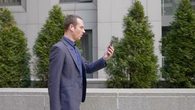 A young businessman walk with wireless earphones in his ears and talking on a video call on smartphone. A guy in a suit speaking on a videoconference. 4K footage