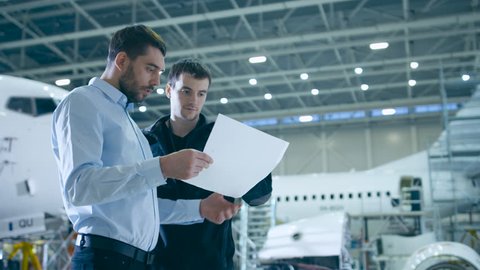 Aircraft Maintenance Mechanic and Chief Engineer Have Discussion, Consult Blueprints While Standing in a Big Airplane Development Facility. They Analyze, Inspect, Develop and Design Airplanes