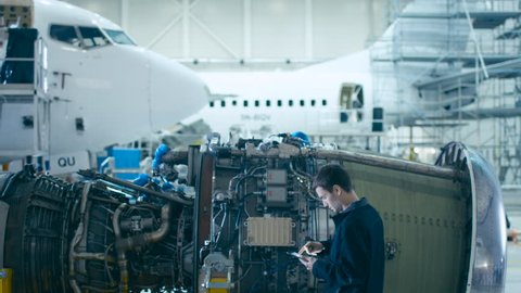 Aircraft Maintenance Mechanic Uses Digital Tablet Computer to Analyze, Inspect and Work on Airplane Jet Engine in Hangar. Optimizing Work, Efficiency and Safe Work of Technology