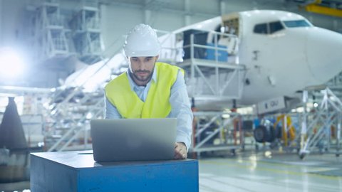 Aircraft Development Engineer wearing Safety Vest and Hardhat Uses Laptop and Blueprints to Analyze, Inspect and Work on Airplane Design in Airplane Design Facility