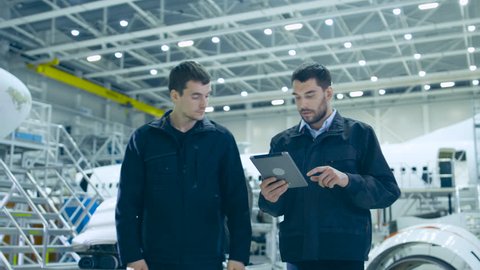 Team of Aircraft Mechanics and Engineers Waking Through Airplane Maintenance, Design and Development Facility. Professionals Consulting Digital Tablet Computer and Talking
