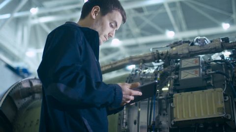 Aircraft Maintenance Mechanic Uses Digital Tablet Computer to Analyze, Inspect and Work on  Airplane Jet Engine in Hangar. Optimizing Work, Efficiency and Safe Work of Technology. 