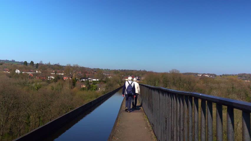 An elderly couple with grey hair walk across the famous Pontcysyllte Aqueduct on the Llangollen canal route in the beautiful Welsh countryside