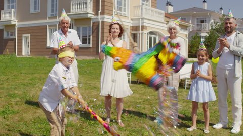 Happy multigenerational family in party hats clapping hands and smiling while watching little boy with blindfold hitting piñata with bat at birthday celebration outdoors on summer day