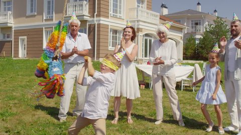 Little boy breaking piñata with bat and joyous family running to him and start picking up treats from grass at birthday celebration outdoors on summer day