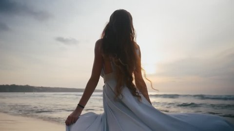 Beautiful young long-haired girl in a free romantic dress walks barefoot on the sea beach leaves traces on the background of the evening sunset and calm sea