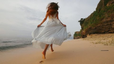 Beautiful young slim girl model runs along the beach in a long evening dress on the background of the sea and sunset and rocks
