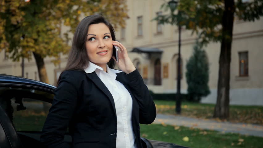 Attractive brunette businesswoman talking on mobile phone with client leaned on car