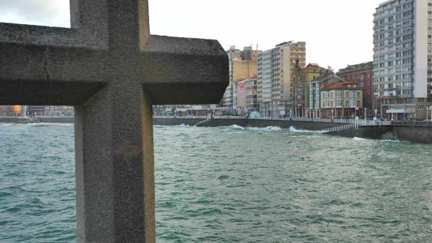 The skyline of the city of Gijon (Asturias, Spain), seen through a big stone cross; high buildings facing the sea, waves shaking the water.