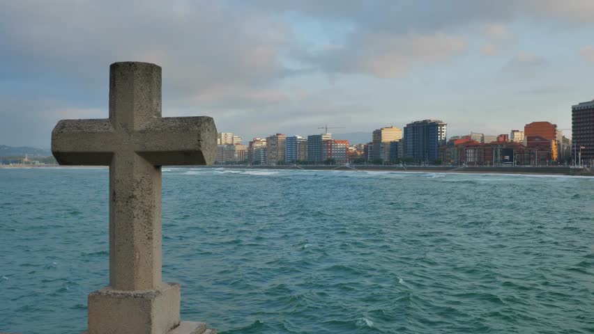 A huge stone cross facing the skyline of the city of Gijon (Asturias, Spain). High buildings on the sea, waves shaking the blue water.