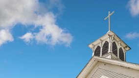One minute looping video footage features white clouds moving over a blue sky above an old bell tower, topped with a cross, on a small rural church painted white. Great text space!
