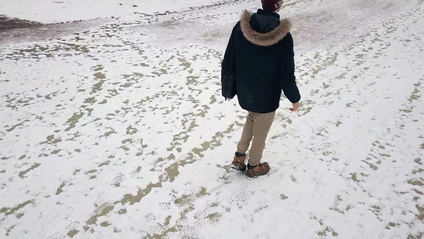 Rising drone footage behind a man walking down a snowy school track in winter gear with a bag.
