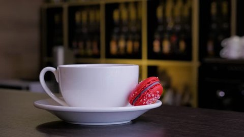 Female hand puts cup of tea with red macaroon on wooden table in cafe